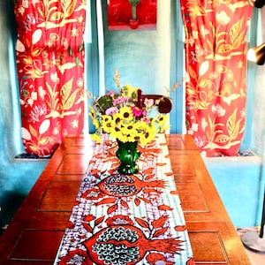 Puede incluir: Una mesa de madera con un camino de mesa colorido con un diseño de granada. Un jarrón verde contiene girasoles y otras flores. Cortinas rojas con estampados florales enmarcan la escena, con una pintura de cactus en el fondo.