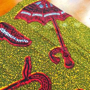 Puede incluir: Tela amarilla con un patrón de espiral verde y diseños rojos y negros. El diseño presenta un paraguas con escritura árabe y una forma curva con un mango rojo y negro.