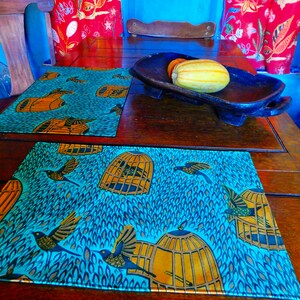 Puede incluir: Dos manteles individuales azules con un patrón de pájaros y jaulas sobre una mesa de madera. Los manteles individuales están hechos de una tela con fondo azul y un patrón de pájaros y jaulas amarillos, naranjas y marrones. La mesa está hecha de madera oscura y tiene un acabado natural.