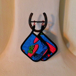 May include: Two blue and red pot holders with a black trim hang from a horseshoe-shaped hook on a white wall.