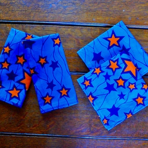 Puede incluir: Cuatro servilletas de tela azul con un patrón de estrellas blancas y naranjas. Las servilletas están dobladas y dispuestas sobre una superficie de madera.