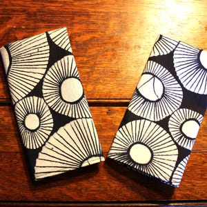 May include: Two folded fabric napkins with a black and white circular pattern, resembling stylized sunbursts. The napkins are placed on a dark wooden surface, showcasing the contrasting colours and design.