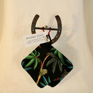 May include: A black kitchen cloth with a green and blue botanical pattern hangs from a rustic horseshoe. A white tag with the words "Kitchen Cloth" is attached to the cloth.