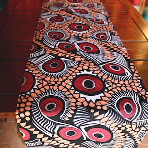 Puede incluir: Un camino de mesa estampado con un diseño repetitivo de grandes formas estilizadas en forma de ojo en rojo, negro, blanco y naranja. El camino de mesa se muestra sobre una mesa de madera oscura, mostrando sus colores vibrantes y su intrincado patrón.