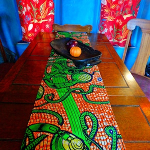 Puede incluir: Una mesa de comedor de madera con un camino de mesa naranja y verde colorido con un patrón repetido de auriculares. Dos frutas naranjas y una fruta morada están en una bandeja de madera en el camino de mesa.