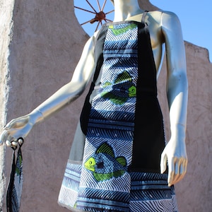 Puede incluir: Un delantal con un estampado de peces en azul, verde y blanco. El delantal tiene un panel negro y un accesorio a juego con estampado de peces. El delantal se exhibe en un maniquí.