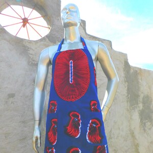 Puede incluir: Un delantal azul con un diseño circular rojo y blanco en el pecho y un patrón de figuras rojas con forma de gallo. El delantal tiene cuello halter y se muestra en un maniquí.