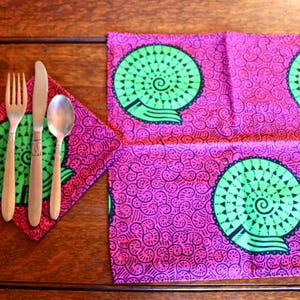 Puede incluir: Una servilleta de tela cuadrada y un mantel individual rectangular con un fondo magenta vibrante, decorado con un patrón repetitivo de espirales negras y grandes motivos circulares verdes. Un tenedor, un cuchillo y una cuchara de plata descansan sobre la servilleta.