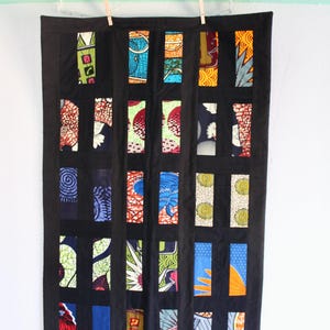 May include: A black quilt with rectangular panels of colorful African fabric. The quilt is hung on a white wall with wooden clothespins. The fabric panels feature various patterns and colors, including blue, green, red, and yellow.
