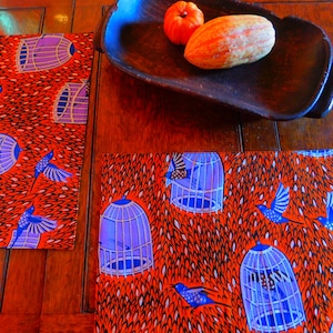 Puede incluir: Dos manteles individuales de color naranja y azul con un diseño de pájaros en jaulas. Los manteles individuales están sobre una mesa de madera.