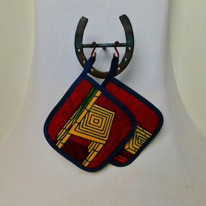 May include: Two red, yellow, and green patterned pot holders with blue trim hanging on a metal horseshoe hook.