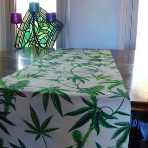 May include: A white table runner with a green marijuana leaf pattern. The runner is on a wooden table.