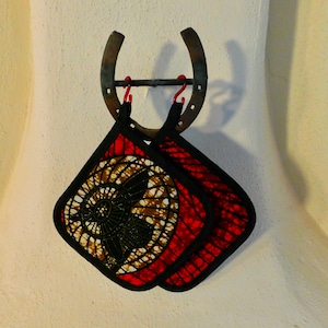 May include: Two red and black pot holders with a brown and white patterned design hang from a horseshoe on a white wall.