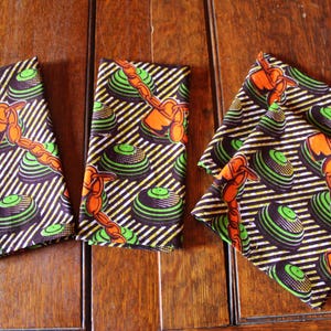 Puede incluir: Un conjunto de servilletas de tela estampadas. Las servilletas presentan un diseño repetido de formas circulares verdes y negras con patrones anaranjados en forma de cadena, sobre un fondo de rayas diagonales blancas y negras.