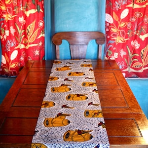 Puede incluir: Una mesa de madera con un camino de mesa estampado. El camino de mesa presenta un diseño repetido de objetos amarillos y pájaros sobre un fondo blanco y azul. Cortinas florales rojas y una pintura de cactus en el fondo.