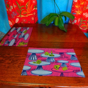 Puede incluir: Dos manteles individuales de color rosa y azul sobre una mesa de madera. Los manteles individuales presentan un diseño de setas estilizadas y tazas. La mesa está puesta con una orquídea blanca en una maceta verde.
