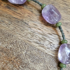 May include: A close-up of a necklace featuring large, polished amethyst beads interspersed with smaller, round green beads. The beads are strung on a dark gray cord, creating a striking contrast against the wooden background.