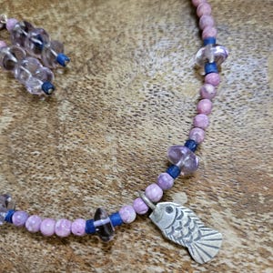 May include: A handmade necklace and earring set featuring a silver fish pendant. The necklace is made of pink and purple beads, blue spacers, and clear crystal beads. The earrings have a similar design. The jewelry is set against a wooden background.