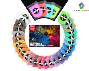 Puede incluir: Un conjunto de 30 paquetes de polvo de mica de colores vibrantes dispuestos en un patrón circular alrededor de una caja central etiquetada como Craft Resin Premium Quality Natural Mica Powder. Cada paquete de plástico contiene un pigmento distinto.