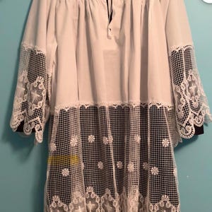 May include: White tunic with long sleeves and a square neckline. The sleeves and lower section feature intricate white lace detailing over black fabric. A black ribbon ties at the neckline. The tunic is likely for religious ceremonies.
