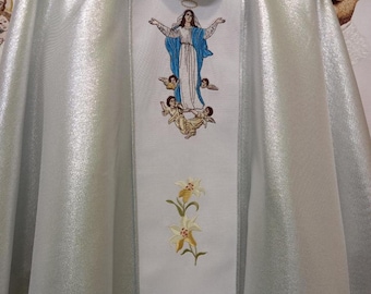 May include: A white liturgical vestment with a shimmering, silvery sheen. The front features an embroidered design of a woman in a blue robe with angels, and yellow lilies. The vestment is likely used in religious ceremonies.