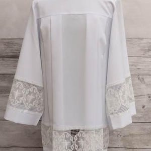 May include: A white liturgical alb with long sleeves, displayed on a mannequin. The alb features a square neckline and is adorned with intricate lace detailing at the cuffs and hem. The lace includes cross motifs.