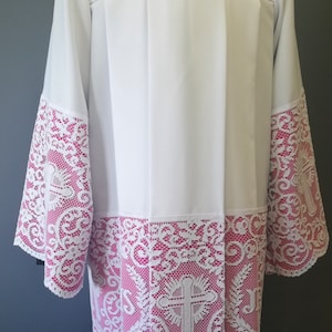 May include: White liturgical vestment with pink lace trim. The lace features a floral design with a cross in the centre.