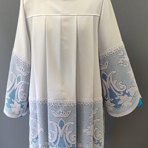 May include: White liturgical vestment with lace trim. The lace is white with a blue background and features a floral design.
