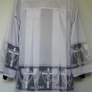 May include: White liturgical alb with a lace trim featuring a repeating pattern of a heart with a cross and grapes, and the letters "IHS".