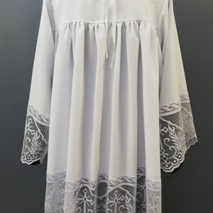 May include: White liturgical alb with lace trim. The lace features a floral design with crosses.