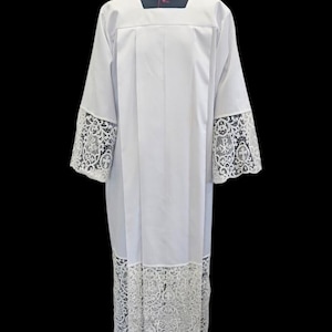 Könnte beinhalten: Ein weißes liturgisches Albe mit quadratischem Ausschnitt, dunkelgrauem Kragen und rotem Zierstreifen. Das Kleidungsstück ist an den Ärmelabschlüssen und am unteren Saum mit breiten Spitzenbesätzen versehen, die florale und religiöse Motive zeigen.