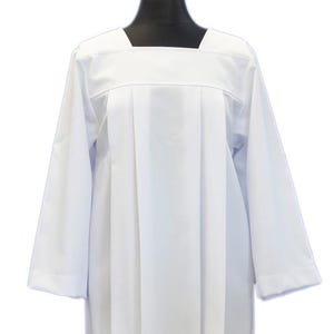 May include: A white, long-sleeved liturgical alb with a square neckline. The garment features pleats down the front and is displayed on a black mannequin head. The sleeves end at the wrists.