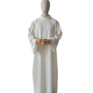 May include: A cream-colored, long-sleeved garment with a high, rounded collar. The garment is displayed on a mannequin with wooden hands clasped together. The fabric appears to be a lightweight material.