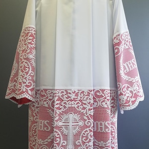 May include: A white liturgical alb featuring wide sleeves and a bottom hem adorned with intricate white lace overlays. The lace pattern includes repeating JHS monograms and central cross motifs set against a vibrant red fabric background.