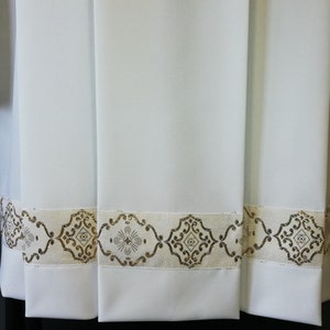 May include: White fabric with a gold and white trim. The trim has a repeating pattern of a diamond shape with a cross inside.