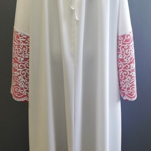 May include: White liturgical alb with red lace trim around the sleeves and bottom hem. The lace features a floral design. The alb has a gathered neckline and a tie at the back.