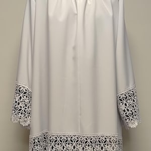May include: A white liturgical alb featuring a high collar with a tie closure at the neck. The garment has long sleeves and a straight hem, both of which are decorated with a wide, intricate white floral lace trim that features circular patterns.