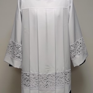 May include: A white liturgical surplice featuring a square neckline and wide sleeves. The garment is adorned with intricate white floral lace trim along the bottom hem and the lower portion of each sleeve, complemented by vertical pleats throughout.