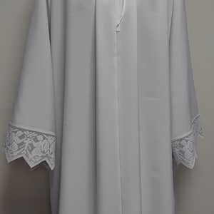 May include: A white liturgical alb featuring long sleeves and a high collar with a tie closure. The garment is decorated with intricate white lace trim along the sleeve cuffs and the bottom hem, which displays a repeating floral and cross pattern.