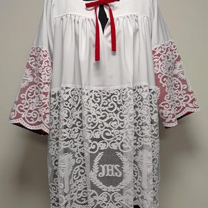 May include: A white liturgical surplice featuring wide sleeves and a lower section adorned with intricate white lace. The lace displays a central JHS monogram surrounded by a wreath and two crosses. A red ribbon tie is centred at the neckline.