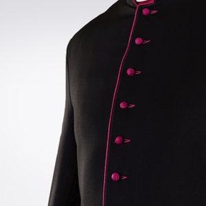 Puede incluir: Casulla negra con ribete y botones rosas. La casulla tiene un cuello mandarín y manga larga.