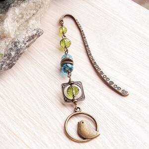 May include: A bronze bookmark with a green and blue glass bead chain and a small bird charm. The bookmark is decorated with a square charm and a circle charm.