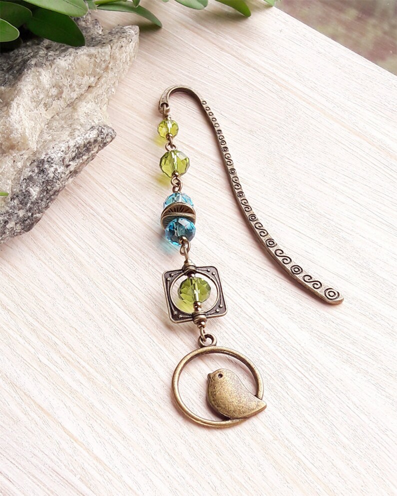 May include: A bronze bookmark with a green and blue glass bead chain and a small bird charm. The bookmark is decorated with a square charm and a circle charm.