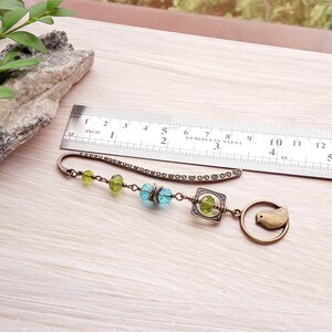 May include: A bronze bookmark with a chain and beads in shades of green and blue. The bookmark features a small bird charm and a square charm with a decorative design.