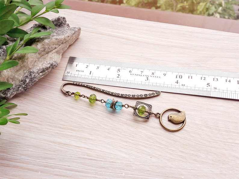 May include: A bronze bookmark with a chain and beads in shades of green and blue. The bookmark features a small bird charm and a square charm with a decorative design.