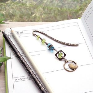 May include: A bronze bookmark with a chain and beads in shades of green and blue. The bookmark features a small bird charm and a square charm with a green glass bead inside. The bookmark is lying on a white page of a planner.