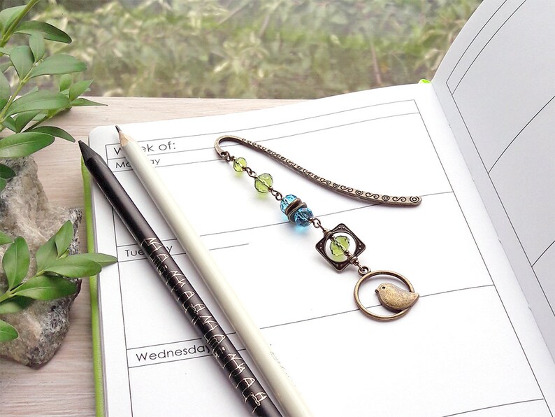 May include: A bronze bookmark with a chain and beads in shades of green and blue. The bookmark features a small bird charm and a square charm with a green glass bead inside. The bookmark is lying on a white page of a planner.