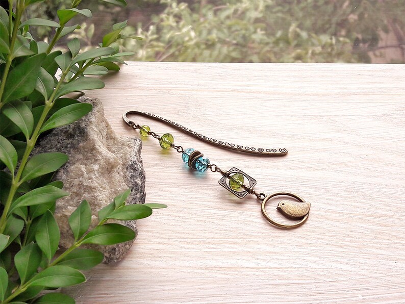 May include: A bronze bookmark with a small bird charm, green and blue glass beads, and a decorative metal charm.