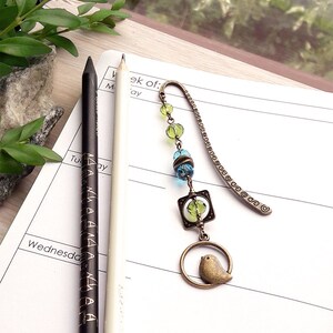 May include: A bronze bookmark with a small bird charm, green and blue glass beads, and a metal chain. The bookmark is lying on a white notebook with the word "Wednesday" written on it.