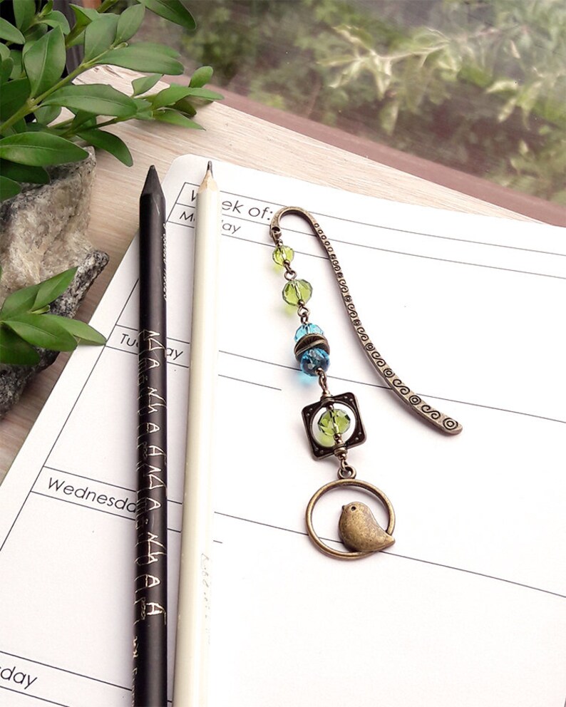 May include: A bronze bookmark with a small bird charm, green and blue glass beads, and a metal chain. The bookmark is lying on a white notebook with the word "Wednesday" written on it.